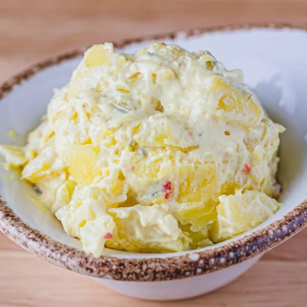 A close-up of a white bowl filled with creamy potato salad, featuring chunks of potato and red specks.